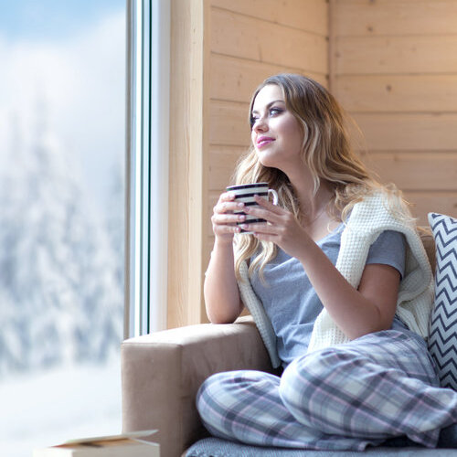 Young beautiful blonde woman with cup of coffee sitting home in living room by the window. Winter snow landscape view. Lazy day off concept